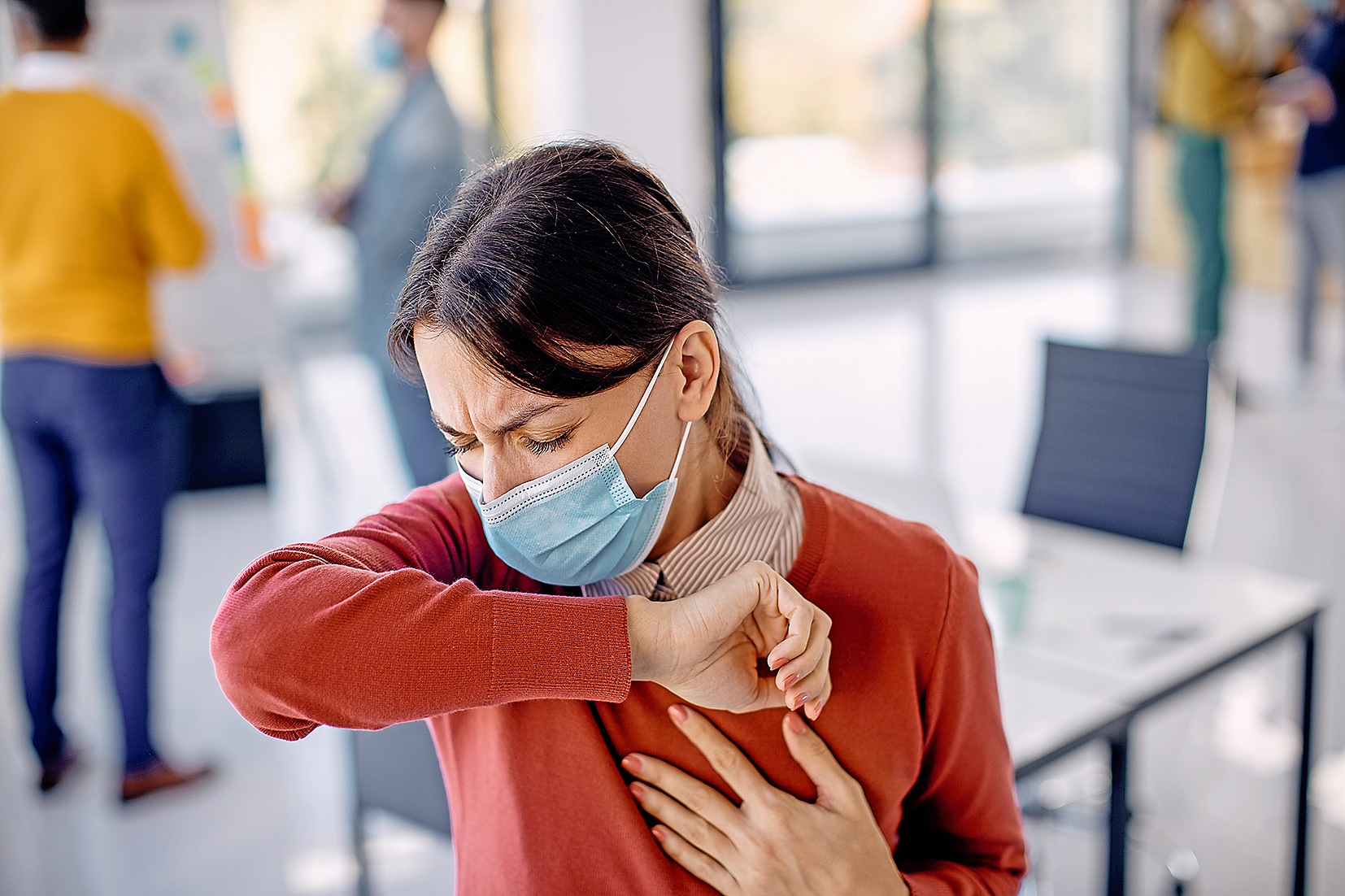 Ill businesswoman sneezing into elbow while wearing face mask in the office.