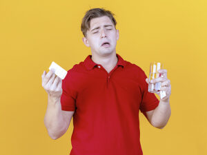 sad young handsome blonde ill man holding pack of medical tablets glass of water and napkin crying with closed eyes isolated on orange background