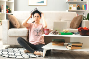 unpleased with closed eyes young girl covered head with notebook sitting on floor behind coffee table in living room